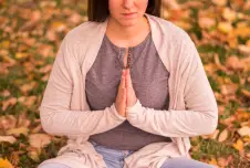 A woman is meditating outdoors with a sad expression, sitting among fallen leaves with her hands in a prayer position